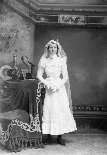 A bride stands in a photography studio for a wedding portrait in a long dress and veil in front of a painted backdrop.