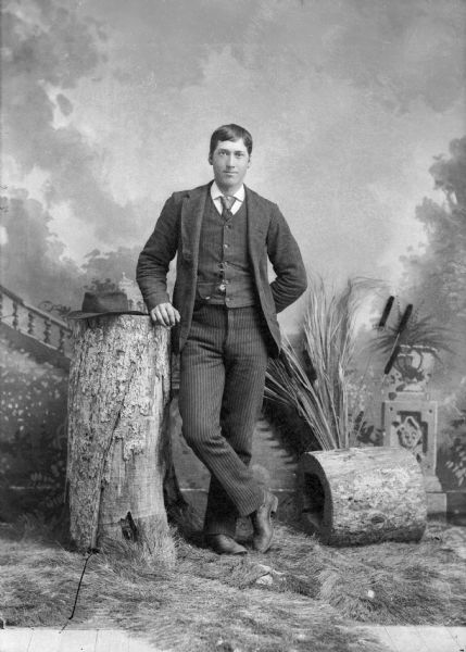 A studio portrait of a young man leaning against a stump in front of a painted backdrop. His hat rests on the top of the stump. He is wearing a suit jacket, vest with watch fob, and trousers.
