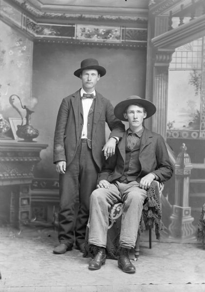 A studio portrait of two young men wearing hats standing in front of a painted backdrop. One young man sits while the other stands and rests his arm on the seated man's shoulder.They are both wearing suit jackets, vests, and trousers.