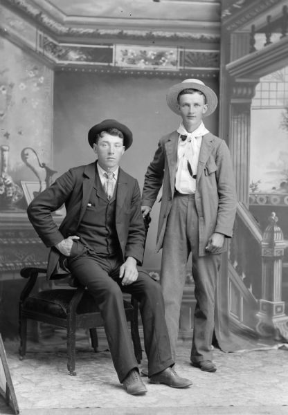 A studio portrait of two young men wearing hats in front of a painted backdrop. One of the young men sits on the arm of a chair while the other stands. They are both wearing He is wearing a suit jackets, neckties, and trousers.
