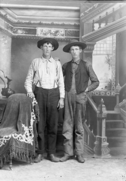Studio portrait of two young brothers, standing side by side. The man on the right has his hand on his brother's shoulder. They are both are wearing hats and standing in front of a painted backdrop. The one on the left is wearing suspenders, and has his hand on a chair draped with a cloth. The man on the right is wearing a vest and overalls.