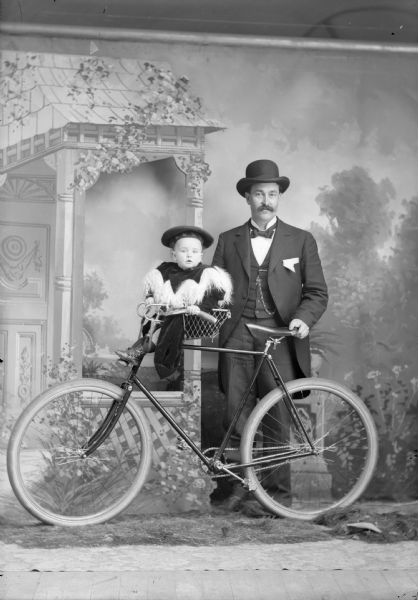 A studio portrait of a man standing and holding a bicycle with an infant in a basket near the handlebars in front of a painted backdrop. The man is wearing a hat, necktie, suit jacket, vest with watch fob, and trousers. The infant is wearing a hat and a coat with a large, fringed collar.