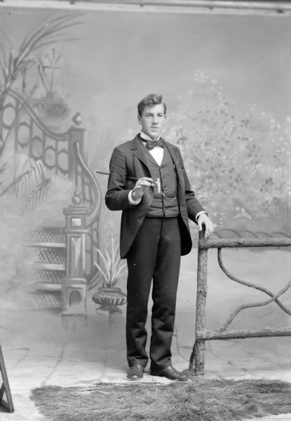 A studio portrait of a well-dressed man in front of a painted backdrop, standing and holding a cane next to a wooden fence. He is wearing a suit jacket, necktie, vest with watch fob, and trousers.