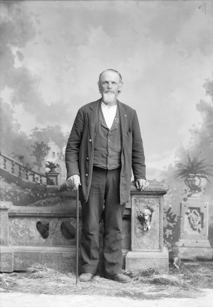 An unidentified elderly man stands with a cane for a studio portrait in front of a painted backdrop. He is wearing a suit jacket, vest, and trousers.