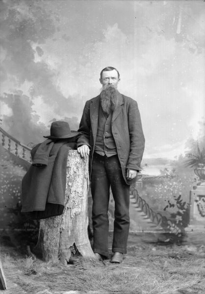A man with a long beard and moustache leans against a stump for a studio photograph in front of a painted backdrop. A coat and hat are on the top of the stump, and he is wearing a suit jacket, vest, and trousers.