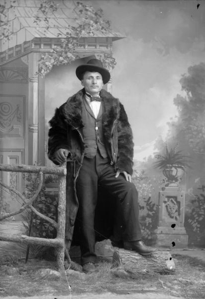Studio portrait of a well-dressed man wearing a fur coat, hat, vest with watch fob, and trousers in front of a painted backdrop. He is posed with his hand on a fence, and his foot on a log.