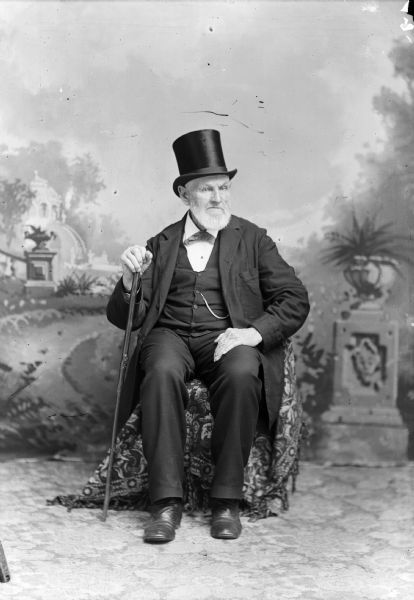 A studio portrait of a sitting man who wears a top hat, suit jacket, vest with watch fob, and trousers. He is holding a cane in front of a painted backdrop.