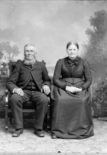 An elderly man and woman sit for a studio portrait in front of a painted backdrop. She is wearing a long, dark dress, and he is wearing a suit jacket over a vest and trousers.