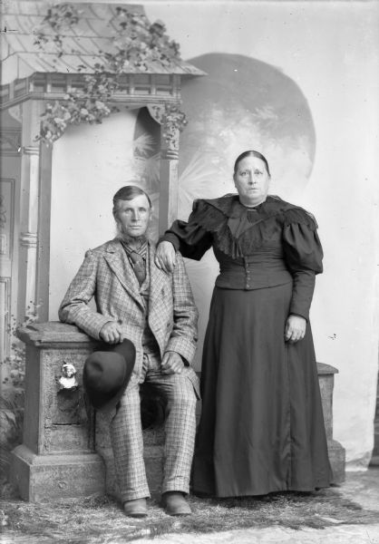 A man and woman pose for a studio portrait in front of a painted backdrop. The man sits on a low fence holding a hat and wears a matching checked suit jacket, vest, and trousers, with a necktie. The woman is in a long, dark dress and stands and rests her hand on the man's shoulder.