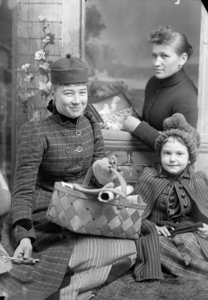 A studio portrait of two women and a young girl, in front of a painted backdrop. The young girl wears a hat and coat and holds a pair of mittens in her lap. The woman next to her wears a hat and coat and holds a picnic basket in her lap and a hand fan in her right hand. The other woman is behind them in a prop window holding a book.