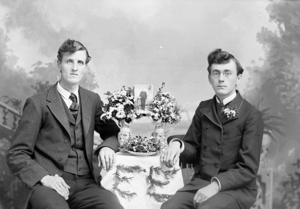 Two men sit in a photography studio and pose with a photograph of a standing man which is displayed between two vases of flowers on a table in front of a painted backdrop. The initials E.E. are spelled out with greenery on the tablecloth. They are wearing suit jackets and trousers, and the man on the right wears eyeglasses and has flowers in his lapel.