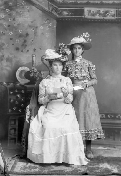 Two young women, one sitting and one standing, pose with a letter for a studio portrait in front of a painted backdrop. Both of the women are well-dressed and wear elaborate hats.