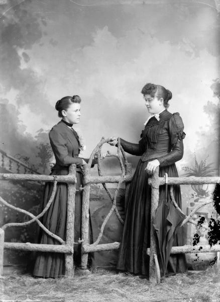 Two women stand facing each other for a studio portrait in front of a painted backdrop. They are wearing long dresses, and are posed near a gate in a prop fence. An umbrella hangs from one of the rails.