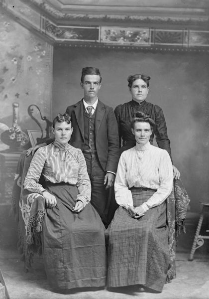 Three women and a young man pose for a studio portrait in front of a painted backdrop. The women are wearing blouses and long skirts. The man is wearing a suit jacket, vest, and trousers.