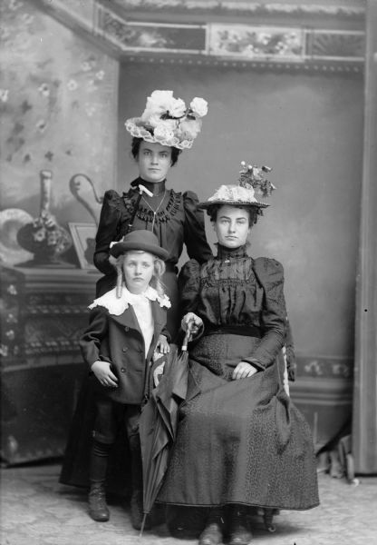 Two women, one sitting holding an umbrella, and the other standing with a cane, pose for a studio portrait with a child (probably a boy) in front of a painted backdrop. Both of the women wear dresses and elaborate hats. The boy wears a hat and a shirt with a lace collar over a suit jacket.