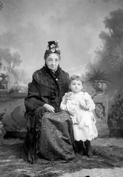 An elderly woman sits with a young girl holding a doll for a studio portrait in front of a painted backdrop. The woman wears an elaborately decorated coat over her dress, and a hat. The girl is wearing a lacy white dress with a sash around the waist.