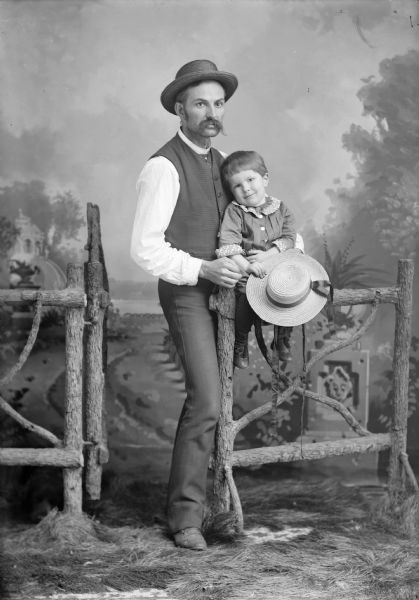 A man stands and holds a small child in front of a painted backdrop on the gate of a prop fence. Identified as Charles Van Schaick and his son. He is wearing a hat, vest, and trousers, and the child is holding a hat.