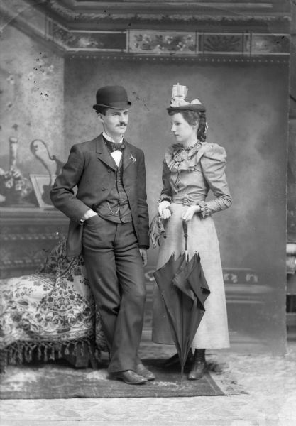A studio portrait of a standing man and woman in front of a painted backdrop. The woman holds a pair of gloves and an umbrella and wears an elaborate hat and ruffled blouse. He is wearing a suit jacket, necktie, vest with watch fob, and trousers, and leans against a chair.