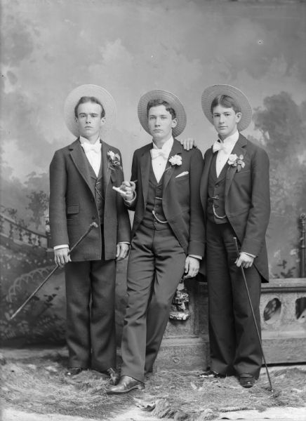 Three men holding canes stand for a studio portrait in front of a painted backdrop near a low fence. The men wear straw hats, white neckties, suit jackets with flowers in the lapels, vests with watch fobs, and trousers.