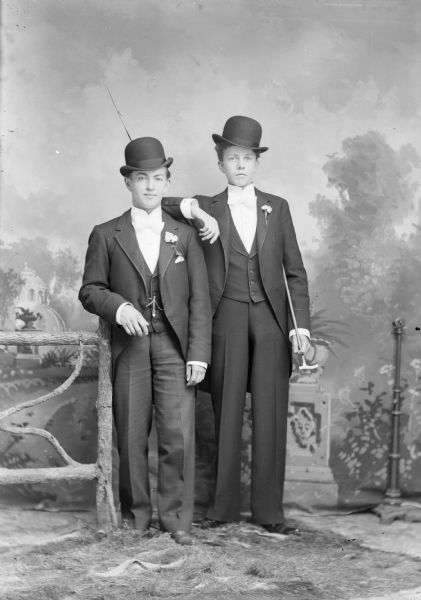 Two well-dressed men wearing hats pose for a studio portrait in front of a painted backdrop near a prop fence. The man on the right holds a cane, and they both wear suit jackets with flowers in the lapel, vests, and trousers.