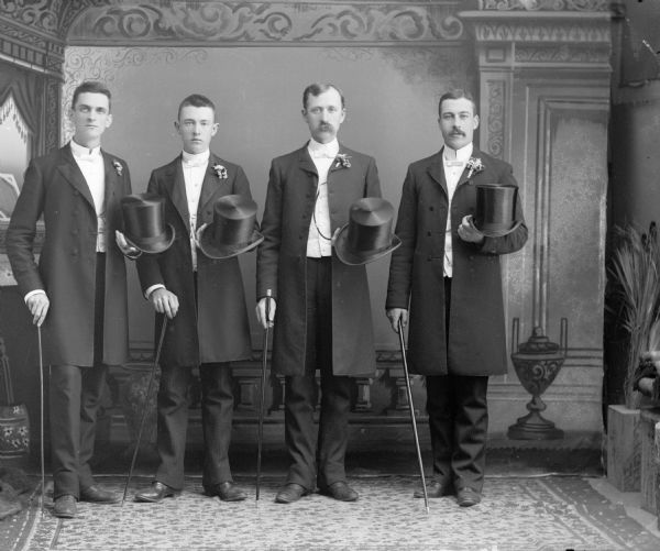 A studio portrait of four standing men in suits holding top hats and canes in front of a painted backdrop. They are all wearing long suit jackets over their trousers, and flowers in their lapels.