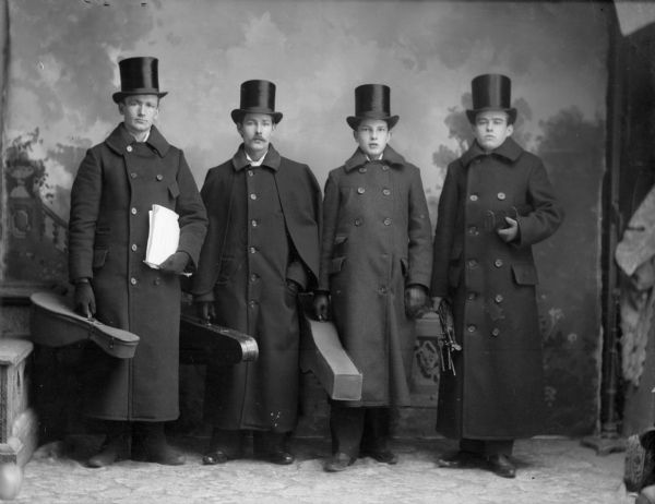 A studio portrait of four well-dressed men carrying instrument cases in front of a painted backdrop. They are all wearing long coats, top hats, and gloves.