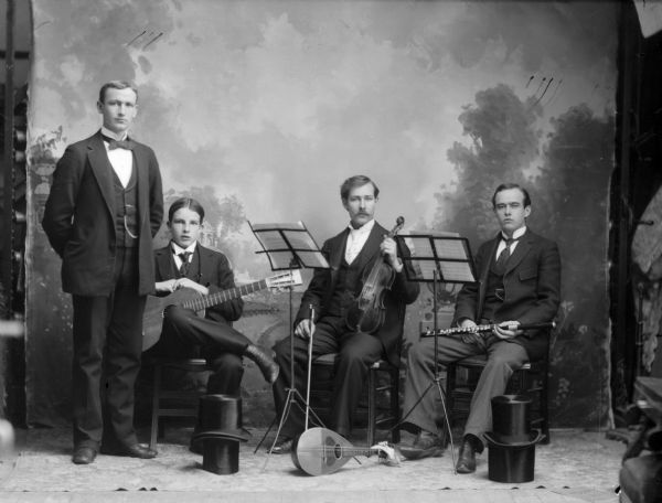 Four musicians in suit jackets, neckties, and trousers, pose with their instruments for a studio portrait in front of a painted backdrop. Their four top hats are stacked together in pairs in front of the musicians on the floor.