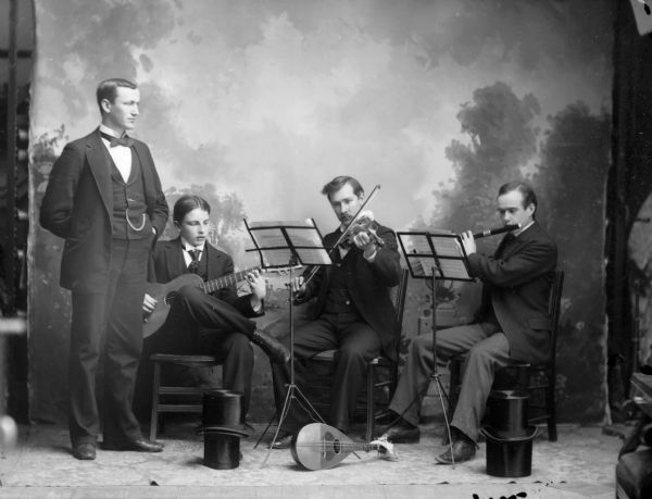 A studio portrait of four men playing musical instruments in front of a painted backdrop. They are wearing suit jackets, vests, neckties, and trousers. The man standing has a watch fob. Four top hats are arranged on the floor in front of the musicians.