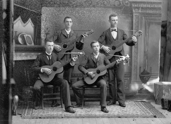 A studio portrait of four men posing with guitars in front of a painted backdrop. They are all wearing suit jackets, vests, neckties, and trousers.