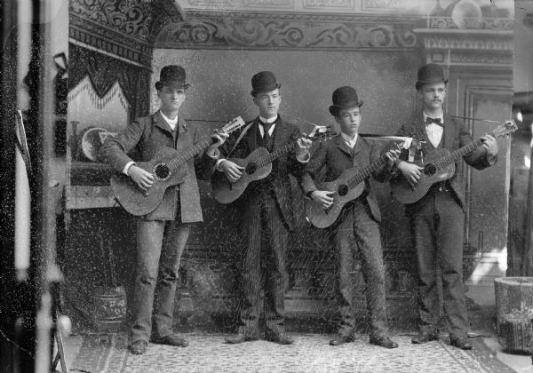 Four musicians stand with their guitars and pose for a studio portrait in front of a painted backdrop. They are all wearing hats, with suit jackets, vests, neckties, and trousers.
