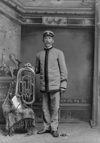 A man wearing a band uniform rests his hand on a tuba and poses for a studio portrait in front of a painted backdrop.