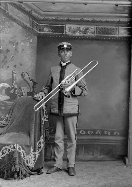 A band member wearing a uniform stands with a trombone, posing for a studio portrait in front of a painted backdrop.