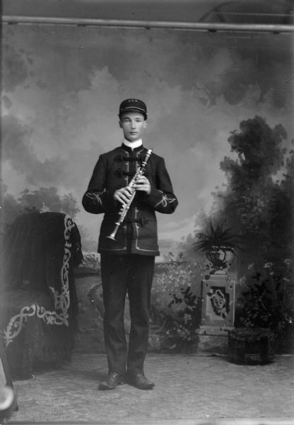 A band member wearing a uniform stands with a clarinet for a studio portrait in front of a painted backdrop.