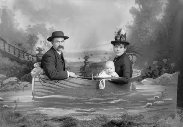 A studio portrait of a couple and an infant sit behind a board painted with a decorative rowboat in front of a painted backdrop. The man and woman are both wearing hats.