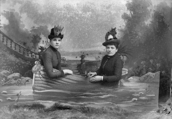 Two women in hats sit behind a board painted with a decorative rowboat for a studio portrait in front of a painted backdrop.