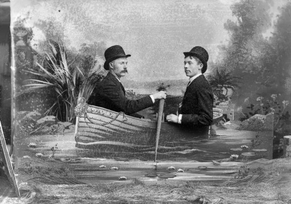 Two men sit behind a board painted with a decorative rowboat and pose for a studio portrait in front of a painted backdrop. Both men are wearing suit jackets and hats.