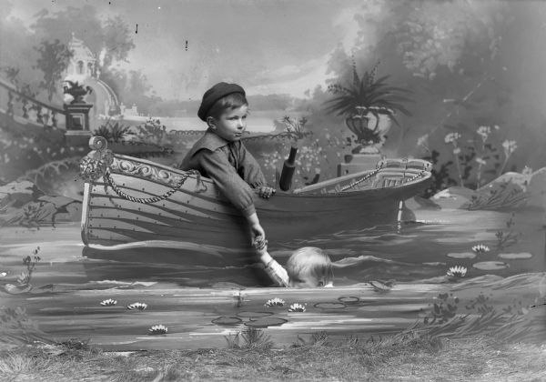 A studio portrait, in front of a painted backdrop, of one boy sitting behind a board painted with a decorative rowboat holding the hand of another boy pretending to be in the water.