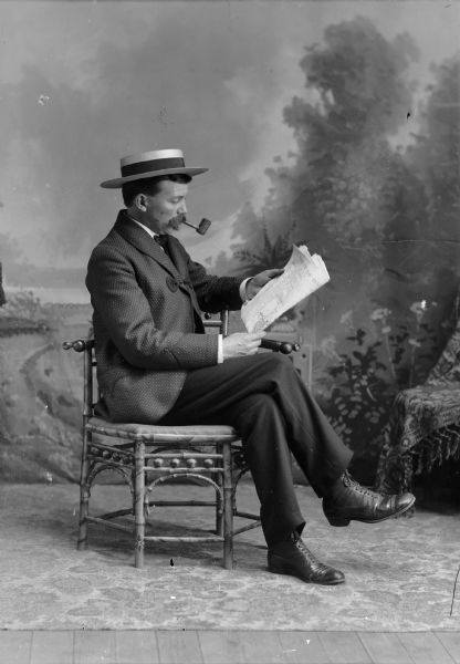 A man sits for a studio portrait in front of a painted backdrop, smoking a corn-cob pipe and reading a Chicago newspaper. He is wearing a hat, suit jacket, and trousers.