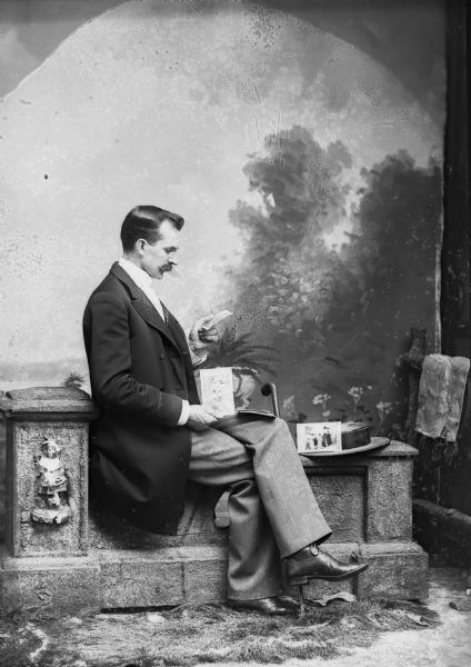 A man, probably Charles J. Van Schaick, wearing a long jacket and trousers, sits on a stone wall in a photography studio and looks at photographs in front of a painted backdrop.