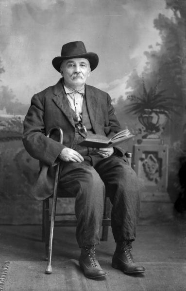 A studio portrait of William Tennant, sitting in a chair wearing a suit jacket and trousers. He is holding a cane and a book in front of a painted backdrop. Mr. Tennant was a magazine and newspaper agent who had two false legs.