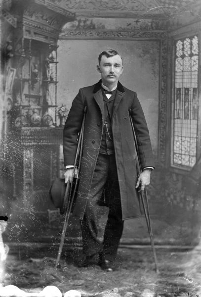 A man stands for a portrait in front of a painted backdrop on crutches. He is wearing a long coat over a vest with a watch fob, and holds a hat in his right hand.