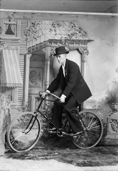 A man poses on a bicycle for a studio portrait in front of a painted backdrop. The man is well-dressed in a hat, suit jacket, and trousers. He wears two unidentifiable medals on his lapel.