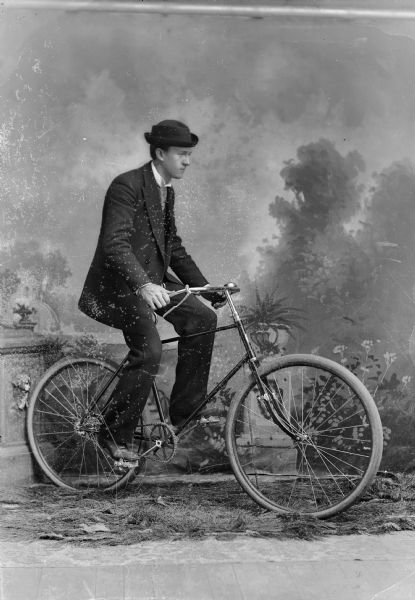 A man sits on a bicycle and poses for a studio portrait in front of a painted backdrop. He is wearing a hat, suit jacket, and trousers.