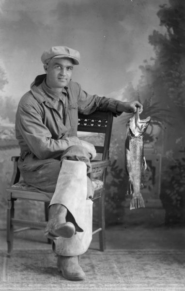 A studio portrait of a sitting man, possibly Earl Davis, holding a trout in front of a painted backdrop. He is wearing a hat, jacket, and long waders folded over beneath the knee.