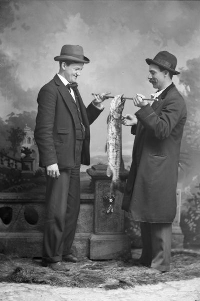 Two men hold a wooden stick and look at a northern pike which is hanging from it.  The two men are well-dressed in hats, suit jackets, trousers, and neckties. They are posed in a photography studio in front of a painted backdrop.