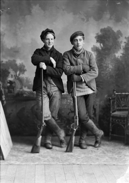 Studio portrait of two young men wearing boots and hats. They are leaning on rifles in front of a painted backdrop.