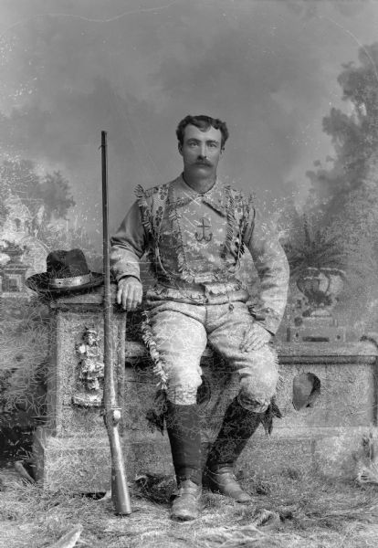 A studio portrait in front of a painted backdrop of a man seated on a stone wall with a rifle resting against the wall to the man's right. The man is dressed in a fringed, leather costume and is possibly a member of a shooting club.