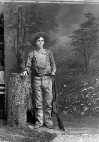 A young man dressed in a fringed, leather costume rests against a stump and holds a rifle for a studio portrait in front of a painted backdrop.