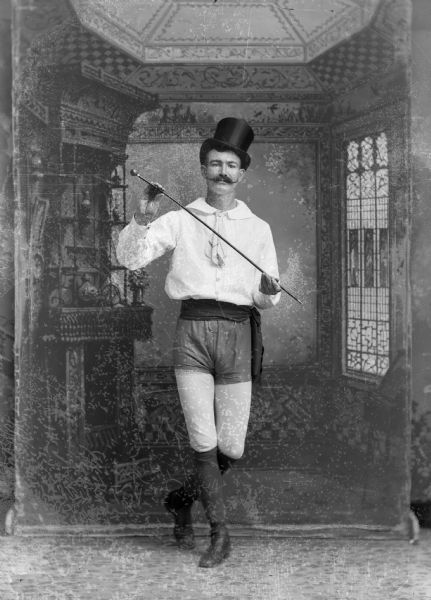 A studio portrait of a standing man in front of a painted backdrop, possibly in a dancing costume with shorts and leggings. He is also wearing a top hat and holding a cane.