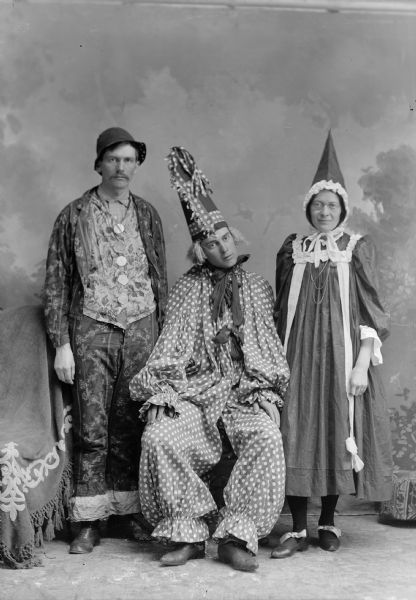 Two men, one seated and one standing, and a standing woman pose for a studio portrait in front of a painted backdrop. All three individuals wear a costume and a hat, possibly dressed as clowns.